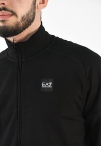 Chaqueta negra de cremallera con cuello alto, textura suave y un prominente parche de logo cuadrado en el pecho.