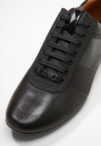 Chaussure en cuir noir présentant des textures lisses et perforées, un bout arrondi, un design classique à lacets, et un logo en métal sur le côté.