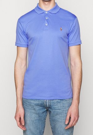 Man draagt een lichtblauw poloshirt met korte mouwen en knopen en vervaagde blauwe spijkerbroek, staand tegen een effen achtergrond.