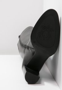 Musta nahkasaapas, jossa on matala korko, kulmaisa pohja eteenpäin osoittavana, koossa 37 ja kohokuvioitu brändilogo pohjassa.