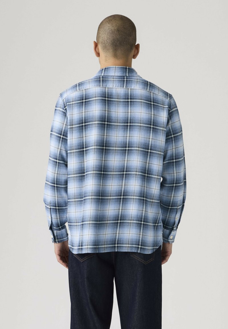 Mand med kort hår iført en blå og hvid ternet langærmet skjorte og mørke jeans, vendt væk mod en ensartet grå baggrund.