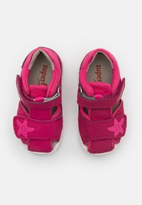 Vaaleanpunaiset lasten kengät, jotka on valmistettu synteettisestä materiaalista. Ominaisuuksiin kuuluvat kaksi säädettävää tarranauhaa ja hymyilevä tähti -kuvio sivussa.