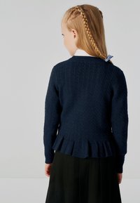 Mornarska pletena cardigan z teksturiranim vzorcem, dolgimi rokavi in ruffled robom. Nositi ga je mogoče preko bele srajce s ovratnikom in plisirane črne krila.