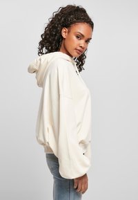 Mlada žena s kovrčavom kosom nosi oversized kremasti hoodie i plave traperice, stoji bočno protiv jednostavne svijetle pozadine.