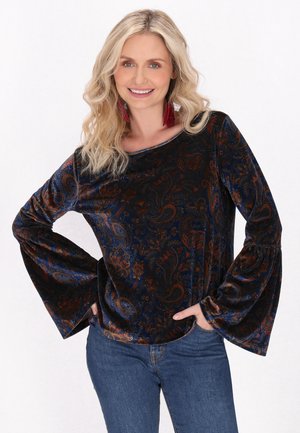 Top in velluto scuro con motivo paisley nei toni di blu, arancio e bordeaux. Presenta maniche a campana e scollatura rotonda. Abbinato a jeans blu.