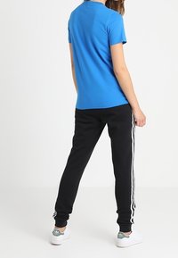 Modrá bavlněná tričko s krátkým rukávem, doplněné černými tepláky se bílými pruhy po stranách. Model otočený zády k fotoaparátu.