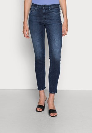 Jeans skinny bleu foncé en denim, avec une taille mi-haute, cinq poches et une texture légèrement délavée. Associés à des sandales noires à enfiler.
