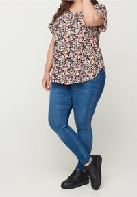 Žena v květované blůze s krátk�ým rukávem, modrých skinny džínech a černých teniskách stojí před jednobarevným pozadím.