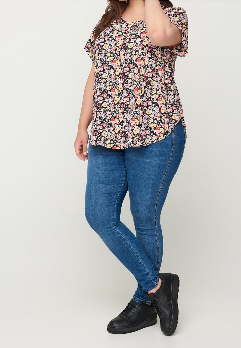 Žena v květované blůze s krátkým rukávem, modrých skinny džínech a černých teniskách stojí před jednobarevným pozadím.