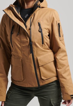 Femme portant une veste à capuche couleur sable avec plusieurs poches zippées noires, associée à un pantalon cargo vert foncé sur un fond uni.