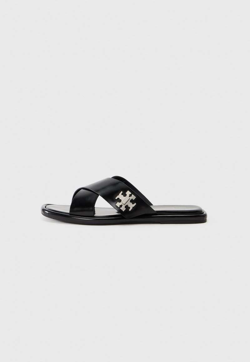 Sandale noire à glissière avec deux brides croisées, fabriquée en cuir lisse. Présente un accent de logo métallique sur le côté et une semelle plate.