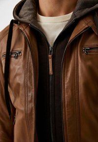 Bruine leren jas met een capuchon, een ritsvoorzijde en zakken aan de zijkanten. In laagjes gedragen met een donkere innerlijke ritskleding en een lichtgekleurde trui eronder.