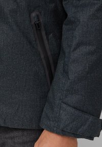 Ciemnoszara tkanina kurtki z teksturowym wykończeniem. Zauważalna boczna kieszeń na zamek błyskawiczny w kolorze czarnym, z gładkim zapięciem. Widoczne detale mankietów.