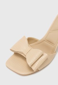 Beige høyhælte sandaler med teksturert materiale, firkantet tå og en stor sløyfe foran, med subtile horisontale linjer.