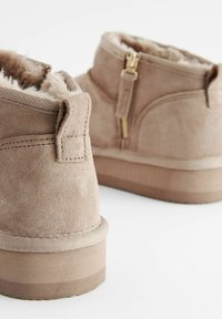 Tan suede ankle boots with faux fur lining, zip closure, and textured rubber sole. Features stitching details and a low heel.