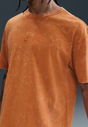 Homme portant un t-shirt Nike orange délavé à manches courtes, montrant une partie des tatouages du bras et les cheveux tressés sur un fond gris.