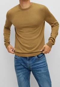 Brązowy sweter z prążkowanej dzianiny, o okrągłym dekolcie i długich rękawach, w połączeniu z niebieskimi dżinsami. Materiał wydaje się być miękki i dopasowany.