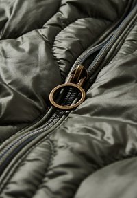 Gros plan d'une doudoune vert foncé partiellement zippée montrant une tirette de fermeture éclair ronde en bronze et une texture de tissu matelassé.