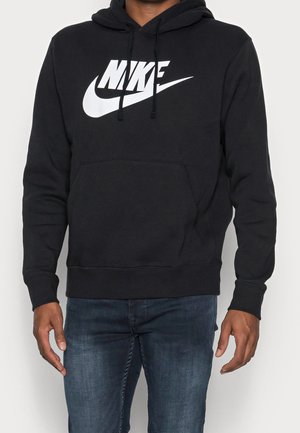 Homme portant un sweat à capuche noir Nike avec logo blanc et jean bleu, debout devant un fond clair uni.