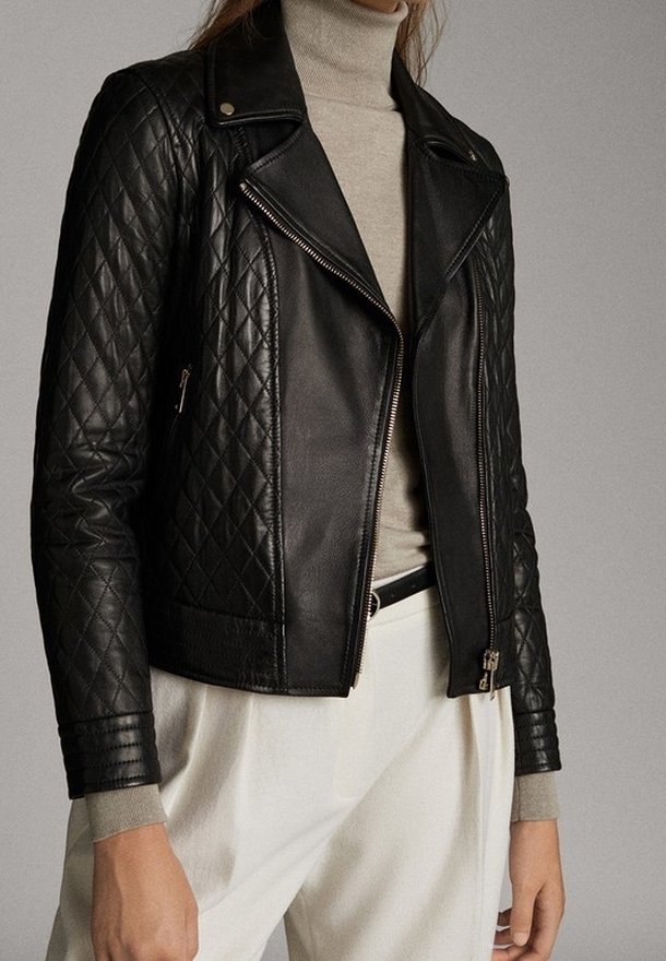 Femme portant une veste moto en cuir matelassé noire sur un col roulé beige et un pantalon blanc avec une ceinture noire.
