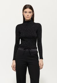 Schwarzes Langarmoberteil mit hohem Kragen, aus strukturiertem, glitzerndem Material. Kombiniert mit schwarzen Hosen mit Kordelzugbund.