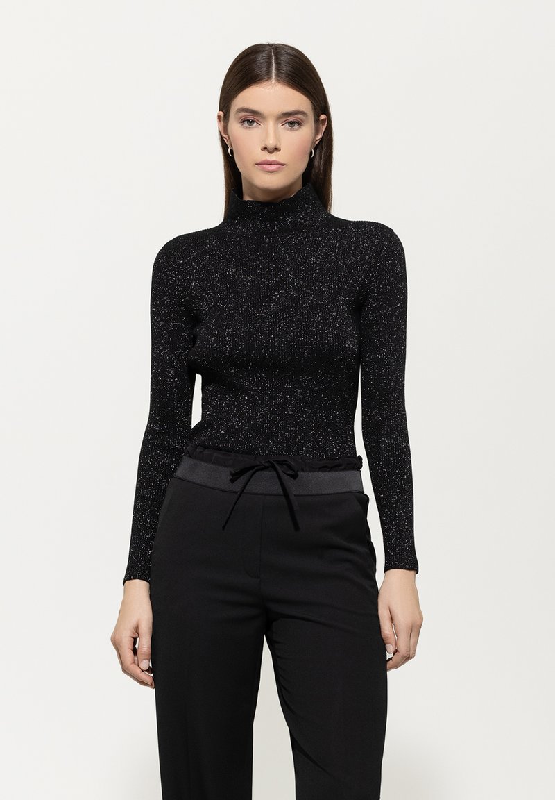 Schwarzes Langarmoberteil mit hohem Kragen, aus strukturiertem, glitzerndem Material. Kombiniert mit schwarzen Hosen mit Kordelzugbund.