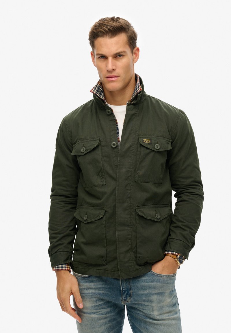 Chaqueta de lona verde con dos bolsillos en el pecho, forro de cuadros visible en el cuello y cremallera frontal. Llevada sobre una camisa ligera y unos vaqueros.