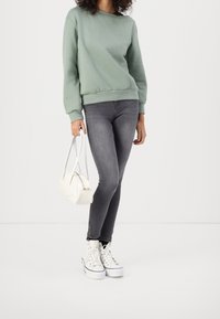 Felpa verde chiaro, jeans skinny grigi, sneakers bianche e una piccola borsa bianca; abbigliamento casual con design e texture semplici.