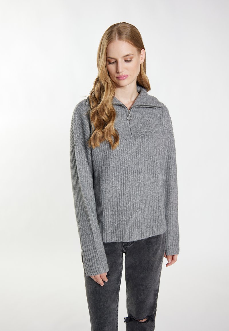 Grauer gerippter Pullover aus weichem Strickstoff, mit einem Viertelreißverschluss, hohem Kragen und langen Ärmeln. Getragen über ausgebeulten schwarzen Jeans.