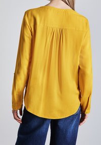 Donna che indossa una blusa gialla senape a maniche lunghe con pieghe sulla schiena e pantaloni in denim blu scuro, in piedi su uno sfondo grigio chiaro uniforme.