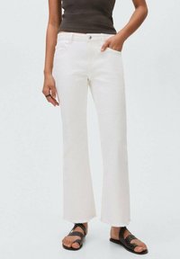 White flared jeans with raw hem, five pockets, button and zipper closure, paired with black strappy sandals on a neutral background.