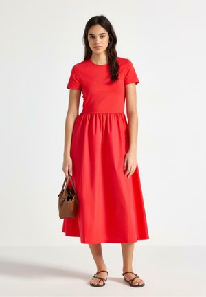 Kvinde iført en rød midi-kjole med korte ærmer, der holder en brun håndtaske og bærer sorte flade sandaler med stropper, ensartet hvid baggrund.