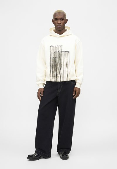 Moški s kratkimi svetlo blond lasmi nosi belo jopa s kapuco, s črnim napisom in vzorcem navpičnih črt, črne široke hlače in črne čevlje, stoji.