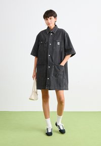 Schwarzes Denim-Hemdkleid mit kragenförmigem Ausschnitt, kurzen Ärmeln, zwei vorderen Taschen und einer Knopfleiste. Präsentiert mit einer weißen Handtasche.