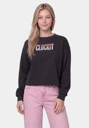Ung kvinde med langt blondt hår iført pink hoop øreringe, en sort sweatshirt med teksten "CLOCKIT", og lyserøde jeans, stående mod hvid baggrund.