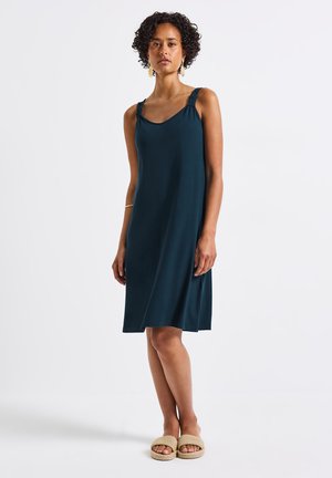 Robe sans manches de couleur bleu-vert foncé avec une coupe décontractée, dotée de bretelles froncées et d'une texture lisse et douce. Portée avec des sandales en osier tressé beiges.