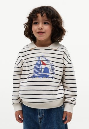 Ungt barn med krøllet hår iført en hvid og navyblå stribet sweater med et sejlskibs-motiv og blå jeans, står mod en hvid baggrund.