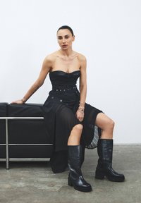 Robe noire en cuir sans bretelles avec un corsage ajusté et une jupe plissée, associée à des bottes noires jusqu'aux genoux avec des semelles épaisses.
