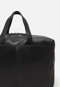 Borsa da viaggio in pelle nera con doppie maniglie, silhouette arrotondata, texture liscia e branding discreto davanti. Chiusura con zip sulla parte superiore.