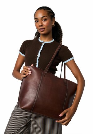 Borsa tote in pelle marrone con trama sottile, linee pulite, doppi manici e cuciture bianche a contrasto. La modella tiene la borsa appoggiata ai pantaloni a righe.
