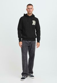 Giovane in piedi, con cappuccio nero con la lettera "B", jeans neri sbiaditi e sneakers bianco e nero, su uno sfondo chiaro e semplice.