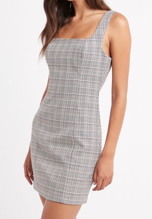 Femme portant une mini-robe sans manches ajustée avec un motif à carreaux noir et blanc et un décolleté carré.