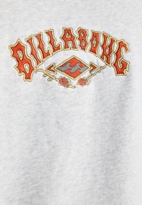 Sudadera gris claro con el texto bordado en rojo y amarillo "BILLABONG" en negrita, acompañado de elementos decorativos y una forma de diamante debajo.