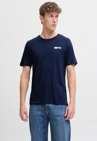 Maglietta in cotone blu navy con maniche corte, colletto rotondo e un logo rettangolare con testo bianco e blu chiaro nell'area sinistra del petto.