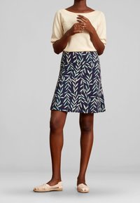 Vaaleanbeige yläosa, tummansininen hame vihreällä lehtikuvioinnilla, liehuva muotoilu, polvipituinen. Kudotut vaaleat kengät viimeistelevät asun.