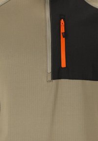 Halvdraget pullover med en beige texturerad kropp och en svart panel vid halsen. Har en orange dragkedjeficka på bröstet.