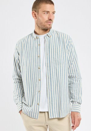 Homme portant une chemise à rayures verticales bleu clair et blanc sur un t-shirt blanc et un pantalon beige, les mains dans les poches, regardant vers la droite.