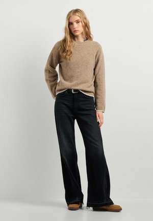 Jonge vrouw met lang blond haar, een beige trui, zwarte wijde jeans en bruine schoenen, staand tegen een effen lichte achtergrond.