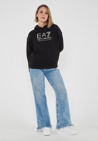 Felpa nera con cappuccio, tasca frontale e logo EA7, abbinata a jeans svasati azzurri e sneakers bianche.