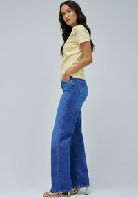Camiseta de algodón amarillo claro con detalles de encaje en los hombros, combinada con pantalones vaqueros anchos de tiro alto en azul y sandalias de tacón beige.